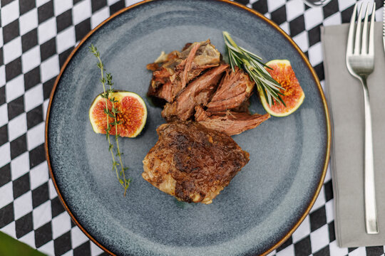 Sliced cooked lamb arranged on a ceramic plate with fresh figs, rosemary, and thyme, served with cutlery on a black-and-white checkered tablecloth