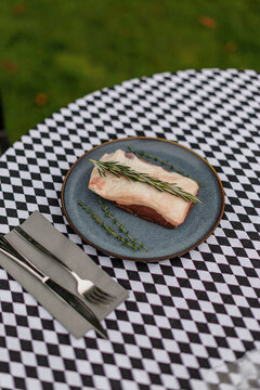 Raw lamb cut topped with fresh rosemary and thyme on a blue ceramic plate, accompanied by fork and knife on a black-and-white checkered tablecloth