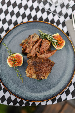 Sliced cooked lamb arranged on a ceramic plate with fresh figs, rosemary, and thyme, served with cutlery on a black-and-white checkered tablecloth