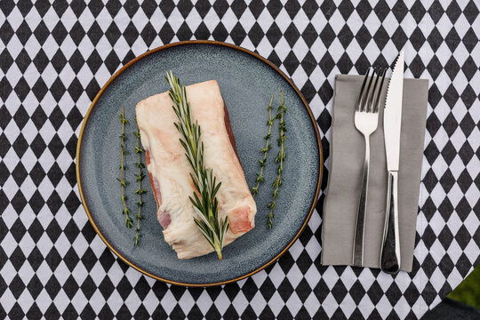 Raw lamb cut garnished with rosemary and thyme on a blue ceramic plate, with fork and knife beside it on a black-and-white patterned tablecloth