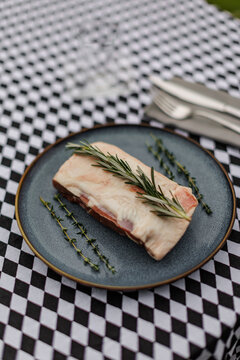Raw lamb meat on a dark ceramic plate, garnished with fresh rosemary and thyme over a black-and-white checkered tablecloth