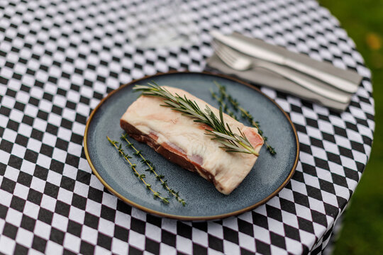 Raw lamb meat on a dark ceramic plate, garnished with fresh rosemary and thyme over a black-and-white checkered tablecloth