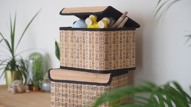Two woven storage boxes stacked on wooden shelf in bright room with green plants background.