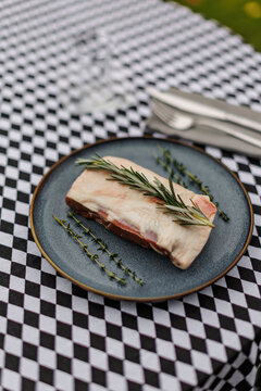 Raw lamb meat on a dark ceramic plate, garnished with fresh rosemary and thyme over a black-and-white checkered tablecloth