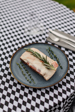 Raw lamb meat on a dark ceramic plate, garnished with fresh rosemary and thyme over a black-and-white checkered tablecloth
