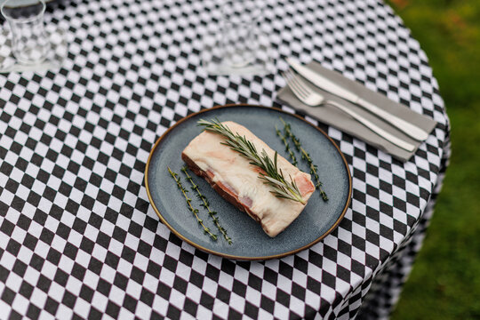 Raw lamb meat on a dark ceramic plate, garnished with fresh rosemary and thyme over a black-and-white checkered tablecloth