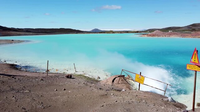 Turquoise geothermal lake near Lake Myvatn, Iceland, with hot water gushing from a fenced outflow and a yellow warning sign on the shore, under a blue sky.