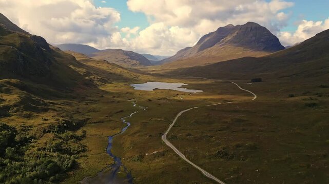 Aerial view of Scottish Highlands with winding paths and serene lakes. Rolling green hills rise under soft clouds. Mountains frame the valley, casting gentle shadows. Stream flows quietly beside fores