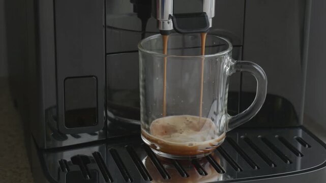 Fresh espresso dripping from coffee machine into glass cup close up.