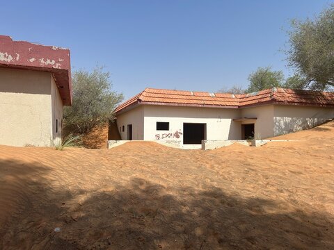 Abandoned village houses partially buried by desert sand dunes under a clear blue sky in an arid landscape with sparse vegetation around