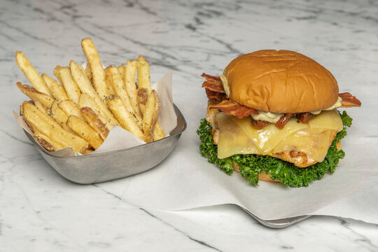 Metal basket of seasoned fries beside a kale-topped chicken sandwich with melted cheese and crispy bacon