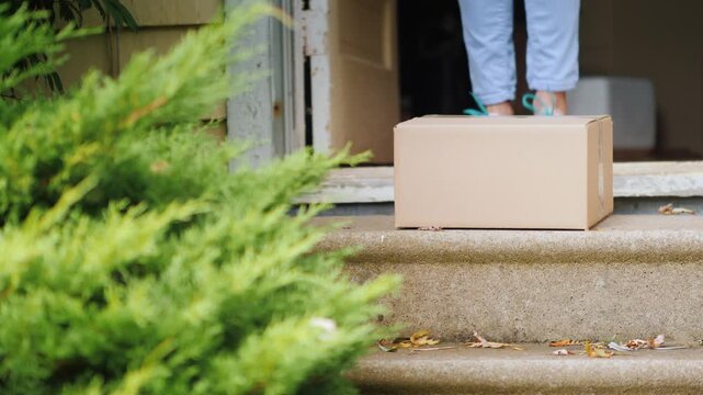 Person picking up cardboard shipping box from front porch step of house.
