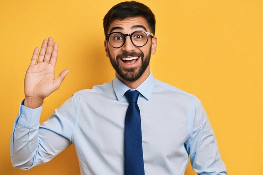 Young handsome businessman with beard, glasses smiles warmly. Happy male manager in blue shirt, tie waves hand for friendly greeting. Confident worker says hello hi to camera on bright yellow
