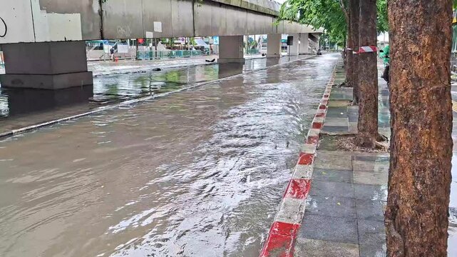 Flooded city street with deep water flowing along sidewalk under elevated skytrain track after heavy monsoon rainstorm in urban area