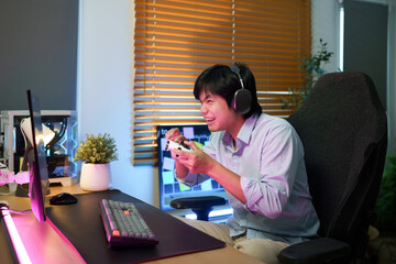 Excited young Asian gamer smiles while holding a controller at a neon lit PC gaming desk, ideal for gaming lifestyle and entertainment concepts © Stella 16