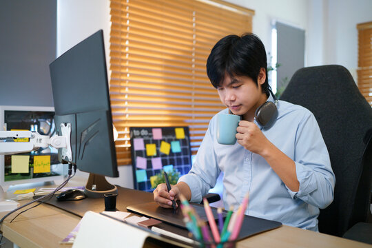 Focused young Asian man sketches on a graphics tablet while sipping coffee, surrounded by monitors and sticky notes, ideal for creative industry concepts