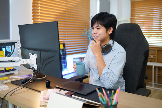 Young Asian male graphic designer holding stylus pen at creative workstation with drawing tablet, monitor, and sticky notes