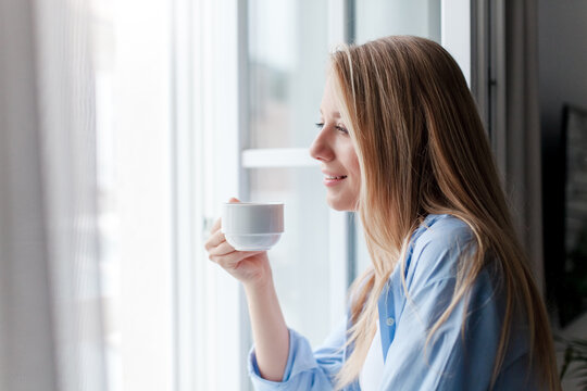 Young woman enjoying morning coffee, calming hot beverage at home. Happy girl relaxing. Slow living, mindfulness, female mental health, recovery. Fresh air, wind from opened window. Airing out room
