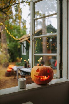 Cozy Halloween window scene: glowing jack-o'-lantern and speckled mug of hot cocoa on rustic windowsill, overlooking a softly lit backyard with string lights and autumn trees &mdash; evoking nostalgia, warm