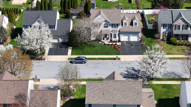 Aerial view of a suburban street featuring detached homes with manicured lawns, blooming spring trees, and a blue car on the road in Lititz, Pennsylvania, United States.