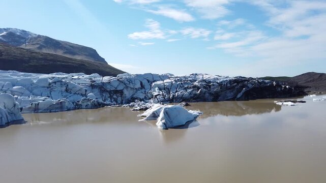 Glacier tongue near Hvannadalshnukur, Iceland, ending in a brown meltwater lake formed by melting snow and ice, dotted with small icebergs, on a sunny day.