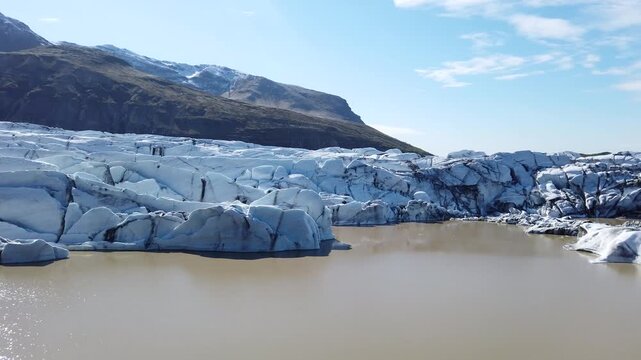 Crevassed outlet glacier of Vatnajokull meeting a muddy meltwater lake below Hvannadalshnukur, Iceland, its ice walls reflected under a clear blue sky.