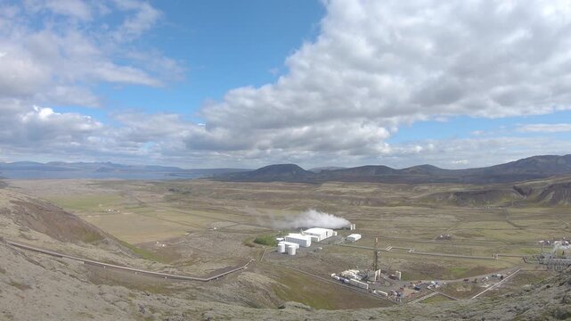 Nesjavellir geothermal power plant releasing steam in a wide volcanic valley, with pipelines crossing the green valley floor.
