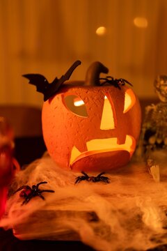 Spooky yet cute Halloween decoration: glowing jack-o'-lantern with bat and spider accents, surrounded by faux cobwebs and plastic spiders on a dimly lit table &mdash; evoking playful horror, nostalgia