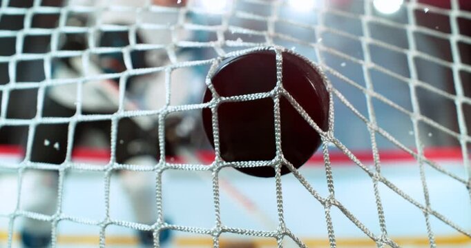 Super slow motion close-up of a hockey puck hitting the goal net as a player scores, captured at 1000 fps, with ice particles and a blurred hockey player on the rink in the background. Speed Ramp.