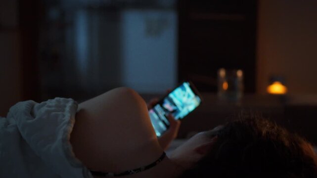 Social media addiction. A young woman lies in bed at night scrolling through her feed on a glowing smartphone. Viewed from behind under a blanket. Theme of screen dependency and sleepless nights