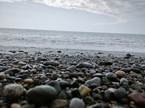 Ground-level perspective of a rocky shoreline with rolling wave. Smooth wet pebbles on the coast under a cloudy sky