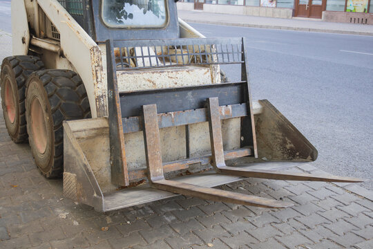 Weathered skid steer loader with a bucket and pallet fork attachment parked beside a city street