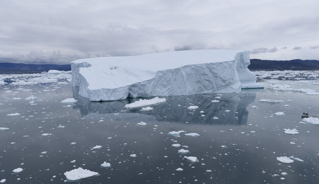 Aerial view of a massive iceberg in the Ilulissat Icefjord, surrounded by ice fragments and calm water under a cloudy sky, with a small boat in Ilulissat, Avannaata Kommunia, Greenland.