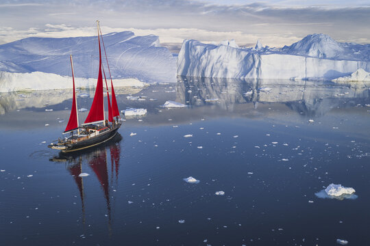 Aerial view of a sailboat with striking red sails navigating through calm dark blue waters filled with massive icebergs in Ilulissat, Avannaata Kommunia, Greenland.