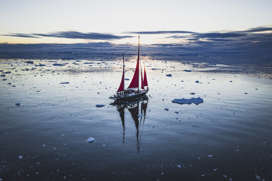 Aerial view of a sailboat with striking red sails navigating through scattered icebergs in the calm waters of the Ilulissat Icefjord at sunset in Ilulissat, Avannaata Municipalit, Greenland.