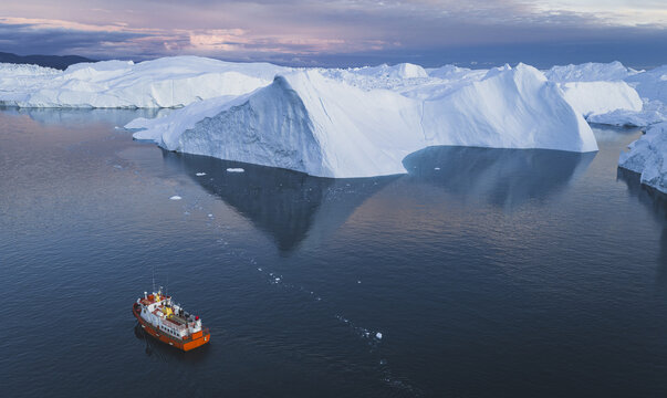 Aerial view of a small orange boat navigating through the massive icebergs of the Ilulissat Icefjord in Ilulissat, Avannaata Municipalit, Greenland.