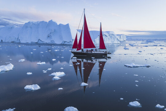 Aerial view of a sailboat with striking red sails navigating through calm waters filled with massive icebergs in Ilulissat, Avannaata Municipalit, Greenland.