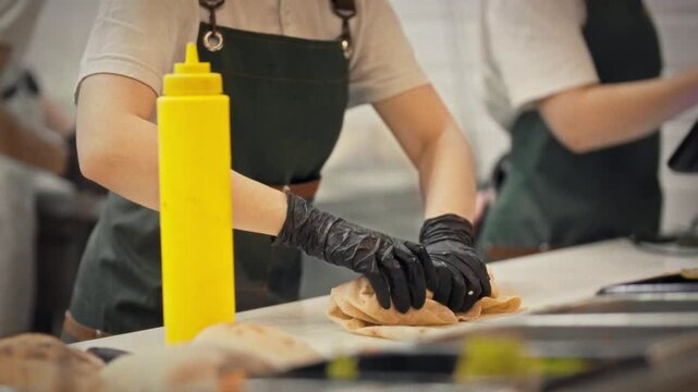 A close-up of a chef wearing an apron and gloves wrapping meat and vegetable filling in lavash. This is a concept for preparing fresh shawarma for takeout at a fast food establishment