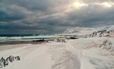 Berlevåg, Norway - 31.03.2019: Snowy arctic coastline with crashing white waves and dark rocks under a heavy, dramatic stormy sky © Andrei