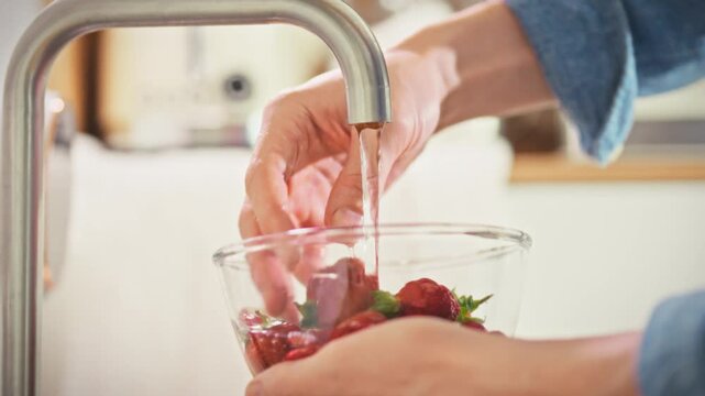 A close-up of a man's hands holding a strawberry under running water in a glass container at home. Maintain a healthy diet and regularly consume fresh seasonal berries