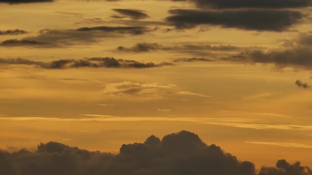 Boiling cumulus clouds morphing and throwing off detached puff that flies up before late breakthrough of intense solar light tints atmospheric layers orange. Dark clouds slide rapidly on foreground.