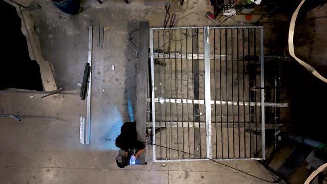 Overhead view of a welder working along a steel gate frame on a workshop floor