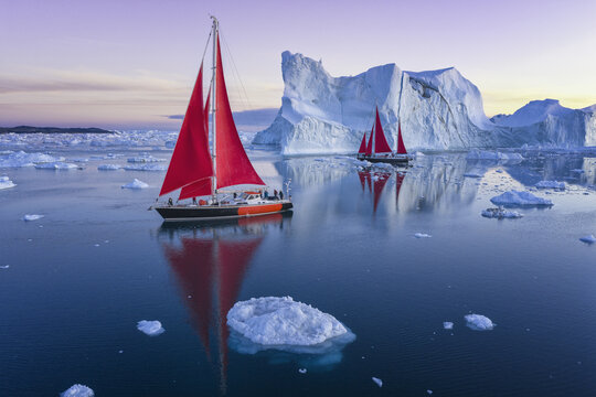 Aerial view of sailboats with striking red sails navigating through massive icebergs in the calm waters of the Ilulissat Icefjord, Ilulissat, Avannaata Municipalit, Greenland.