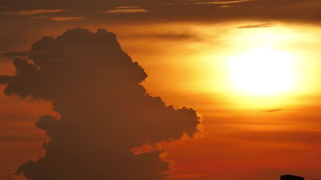 Towering dark cumulus cloud formation resembling giant profile head growing and developing open mouth shape while brilliant setting sun halo moves downward in background. Strange pareidolia cloud.