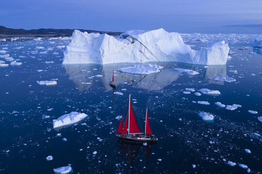 Aerial view of a sailboat with striking red sails navigating through calm, icy waters surrounded by massive icebergs in the Ilulissat Icefjord, Ilulissat, Avannaata, Greenland.