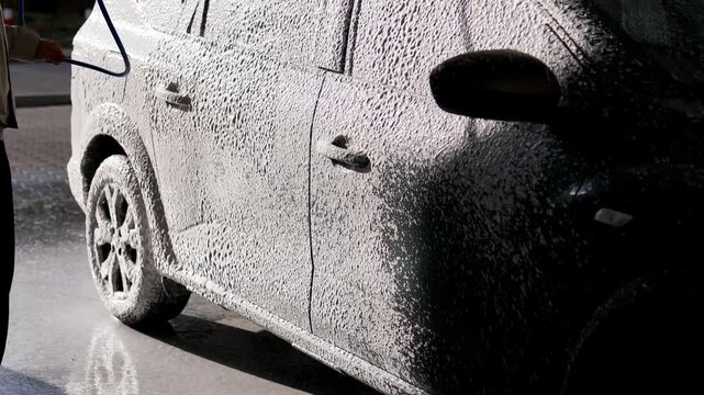 A young man using a professional high-pressure water gun to wash off active soap foam from the body and rear window of a grey crossover SUV car. Action shot at an outdoor self-service carwash station