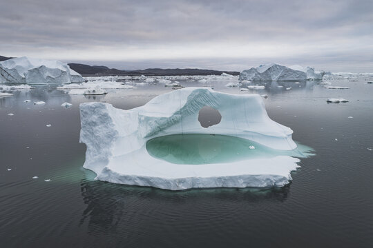 Aerial view of an iceberg with a natural hole and a turquoise meltwater pool floating in the sea near Ilulissat, Avannaata Municipalit, Greenland.