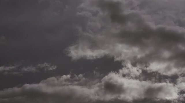Two winds, one sky. A conceptual telephoto time-lapse of atmospheric paradox where opposing forces operate within unified gray canvas. Stratum of dark clouds descends on back, ragged clouds drift up
