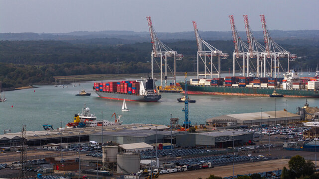 Aerial view of a large container ship navigating the busy port of Southampton with industrial cranes, cargo containers, and small boats in the water, Southampton, England, United Kingdom.