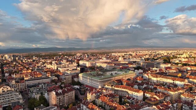A dense European city is illuminated by warm evening light beneath rain clouds and a faint rainbow, viewed from a moving drone.